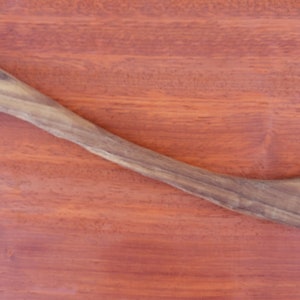 May include: A handcrafted wooden fork and spoon combination tool. The utensil is made of light brown wood and features a fork on one end and a spoon-like scoop with a hook on the other. The tool is set against a reddish-brown wooden surface.