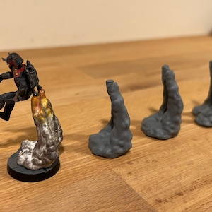 Peut inclure: Une figurine miniature d'une personne en costume noir et rouge avec un jetpack, volant au-dessus d'un effet de fumée gris et blanc. Il y a quatre autres effets de fumée gris et blanc en arrière-plan.