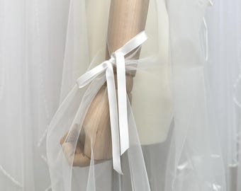 Könnte beinhalten: Ein durchscheinender weißer Stoff ist über einen hölzernen Mannequinarm drapiert. Ein weißes Satinband ist um den Arm gebunden, mit langen Bändern. Der Stoff ist halbtransparent, so dass der Mannequin teilweise sichtbar ist. Der Gesamteindruck ist zart und luftig.