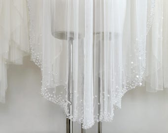 May include: A white wedding veil with a beaded trim, displayed on a mannequin. The veil's sheer fabric is visible, with a detailed beaded edge. The design is elegant and suitable for a bride.