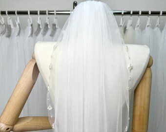 Puede incluir: Un velo de novia blanco con un borde de cuentas. El velo se muestra en un maniquí.