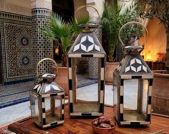 May include: Three decorative lanterns with geometric black and white patterns and gold handles sit on a wooden table. The lanterns are different sizes and have glass panels. A small bowl of dates and a sprig of lavender are also on the table.