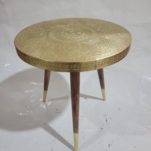 May include: A round, gold-colored side table with an intricate, embossed design on the top. The table has three wooden legs with gold-colored tips.