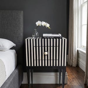 May include: A black and white striped nightstand with two drawers and a round knob. The nightstand has black legs and is topped with a vase of white orchids and two books, one titled "THE ART BOOK".