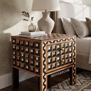 May include: A square wooden nightstand featuring two drawers with a geometric inlay pattern of black, white, and natural wood squares. The frame is accented with a black and white zigzag border and two small gold knobs. The text MOVADECOR is visible.