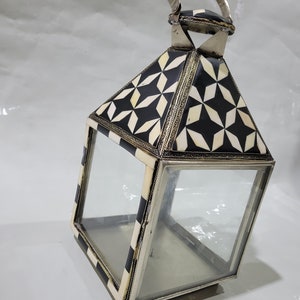 May include: A black and white geometric patterned lantern with a silver handle and glass panels. The lantern is made of bone and metal.