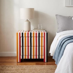 May include: A rectangular bedside table featuring two drawers with a vertical striped pattern in orange, red, black, and white. A beige table lamp with a cylindrical shade and a clear glass tumbler sit on the flat top surface of the furniture.