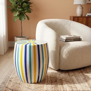 Puede incluir: Una mesa auxiliar con forma de barril que presenta un patrón vibrante de rayas verticales en tonos alternos de amarillo brillante, azul real y verde azulado. La superficie superior muestra un diseño de rayos de sol desde el centro.