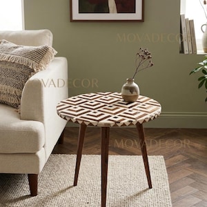 May include: A round side table with a geometric pattern on the tabletop. The table has dark brown wooden legs and a light-colored patterned surface. A small vase with a plant sits on the table. The table is placed on a neutral-colored rug.