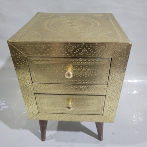 Puede incluir: Una mesita de noche de metal dorado con tres cajones y diseños en relieve intrincados. La mesita de noche tiene patas de madera.