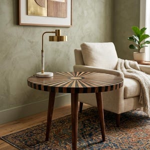 Puede incluir: Una mesa auxiliar redonda de madera con un diseño de rayos de sol en la superficie. La mesa tiene tres patas de color marrón oscuro y una lámpara dorada con base de mármol. La mesa está sobre una alfombra estampada.
