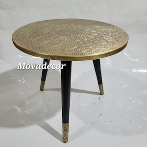 Pode incluir: Uma mesa auxiliar redonda de cor dourada com desenhos gravados intrincados na parte superior. A mesa tem três pernas de madeira preta com detalhes dourados.