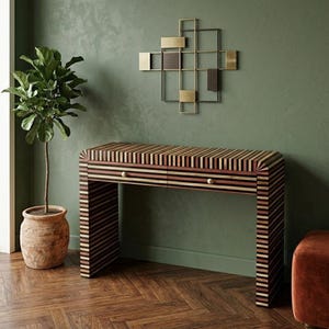 May include: A rectangular console table featuring a multi-colored striped pattern of wood slats in shades of brown, tan, and black. The table has two drawers with small round gold knobs and curved edges. Above it hangs a geometric metal wall sculpture.