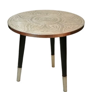 May include: A round silver table with an intricate embossed design on the top. The table has three black legs with gold accents.