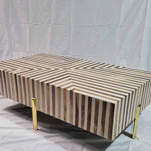 May include: A coffee table with a geometric pattern of white and brown wood stripes. The table has four gold metal legs.