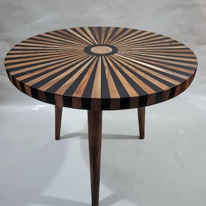 May include: A round wooden table with a black and brown striped top and three dark brown legs. The table top has a sunburst design with a small circle in the center.