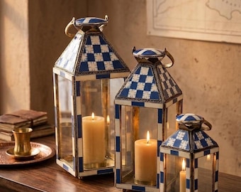 May include: Three decorative lanterns with lit candles. The lanterns have a geometric blue and white pattern on the top and base, with gold-colored metal frames. The lanterns are different sizes and sit on a dark wooden surface.