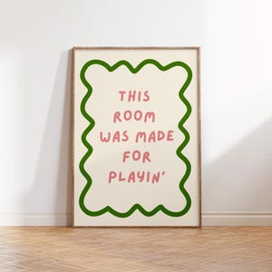 Puede incluir: Una impresión artística enmarcada con el texto "THIS ROOM WAS MADE FOR PLAYIN'" en letras rosas. El texto está centrado en un fondo crema, enmarcado por un borde verde ondulado. El marco es de color marrón claro.