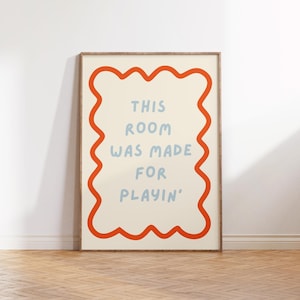 Könnte beinhalten: Ein gerahmter Kunstdruck mit dem Text "THIS ROOM WAS MADE FOR PLAYIN'". Der Text ist hellblau auf cremefarbenem Hintergrund und wird von einem wellenförmigen orangefarbenen Rand umrahmt. Der Rahmen hat eine natürliche Holzfarbe.