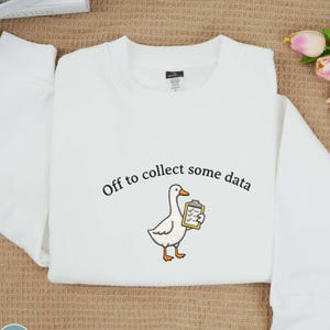 May include: White sweatshirt with the phrase "Off to collect some data" above an embroidered cartoon goose holding a clipboard. The goose has orange feet and beak. The sweatshirt has a crew neck and long sleeves.