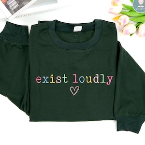 May include: A dark green sweatshirt with the embroidered phrase "exist loudly" in colorful letters and a small heart. The sweatshirt has a crew neck and long sleeves with ribbed cuffs.