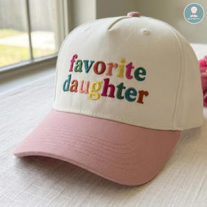 May include: A white and pink baseball cap with the embroidered text "favorite daughter" in colorful letters. The cap has a pink bill and a pink button on top. The hat is a casual accessory.