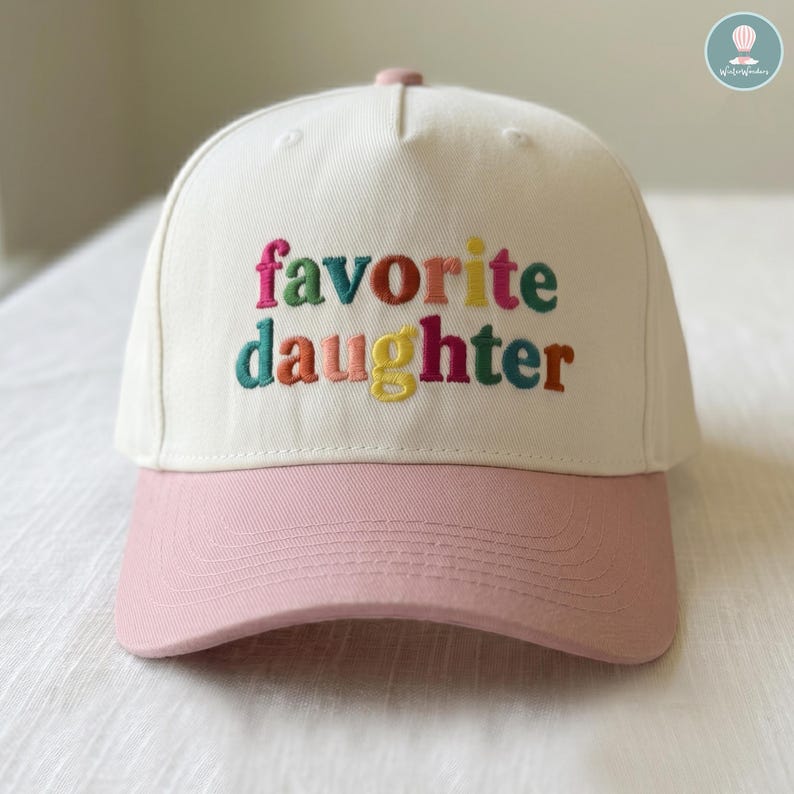 May include: A baseball cap with a white crown and a pink bill. The phrase "favorite daughter" is embroidered in colorful letters across the front. The cap has a pink button on top.
