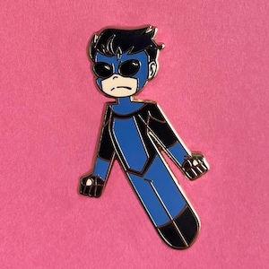 Op de afbeelding: Een pin met een gestileerd superheldenpersonage in een blauw en zwart kostuum met een gouden omtrek. Het personage heeft een masker en staat in een dynamische pose. De pin staat tegen een roze achtergrond, met de tekst "LEATHEROSEART" onderaan.