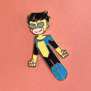 Op de afbeelding: Een metalen speld met een stripfiguur superheld in een dynamische pose. De speld heeft een zwart, geel en blauw kleurenschema, met de gezichts- en kostuumdetails van het personage duidelijk gedefinieerd. De speld staat tegen een perzikkleurige achtergrond. De tekst "LEATHEROSEART" staat onderaan.