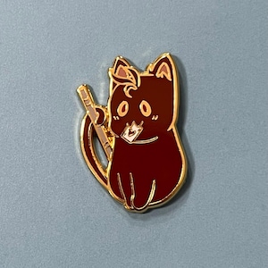 Pode incluir: Um broche de esmalte de gato marrom com contorno dourado. O gato está segurando um pequeno envelope branco na boca e tem um bastão dourado atrás.