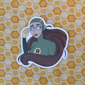 Op de afbeelding: Een sticker met een cartoonillustratie van een persoon met bruin haar, een bril en een groene outfit met een grote letter "R" op de borst. De achtergrond is een geel en wit honingraatpatroon.