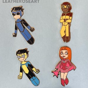 Op de afbeelding: Vier emaille pins met stripfiguren in superheldenposes. De pins hebben goudkleurige metalen randen en tonen personages in blauwe, gele, rode en bruine kostuums. De tekst "LEATHEROSEART" staat linksboven.