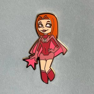 Op de afbeelding: Een speld met een stripheldin met rood haar, een roze cape en een stervormig accessoire. Het personage is gekleed in roze en rode tinten, met een gouden omtrek. De tekst "LEATHEROSEART" staat onderaan.