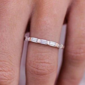 Puede incluir: Un delicado anillo con una serie de piedras preciosas de corte rectangular engastadas en una banda. El diseño del anillo presenta un patrón continuo de piedras preciosas y segmentos metálicos alternos, creando un aspecto refinado y elegante.