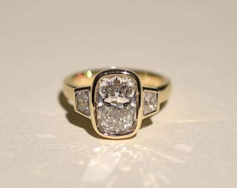 May include: A gold ring featuring a large, rectangular-cut diamond center stone, flanked by two smaller, square-cut diamonds. The diamonds are set in a bezel setting, and the band is a polished gold.