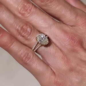 May include: A gold engagement ring featuring a large oval-cut diamond. The central diamond is encircled by a halo of smaller diamonds. The band has a split shank design, also adorned with small diamonds. The ring is shown on a hand.