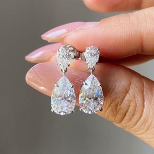 May include: A pair of elegant drop earrings featuring pear-shaped, clear gemstones. The earrings are set in a silver metal, with a smaller gemstone at the top of each earring. The gemstones sparkle, reflecting light. The earrings are held up by a person's hand.