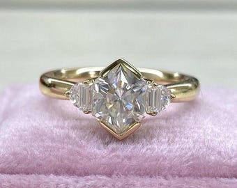 Puede incluir: Un anillo de oro con una gran gema clara y multifacética en el centro, flanqueada por dos gemas rectangulares más pequeñas. El anillo está sobre una superficie de terciopelo rosa, mostrando su intrincado diseño y brillo.