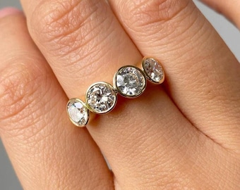 May include: A gold ring featuring four round, clear diamonds set in individual bezels. The diamonds are of varying sizes and sparkle brightly. The ring is worn on a finger, showcasing the jewelry's design and craftsmanship.