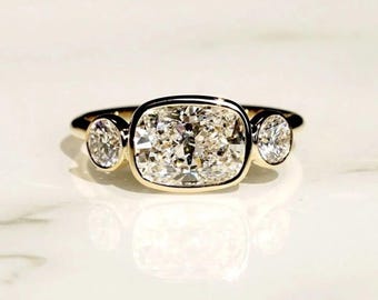 May include: A three-stone ring with a large, square-cut diamond center stone and two smaller round diamonds on either side. The diamonds are set in a gold band, creating a classic and elegant look. The ring is displayed on a white surface.