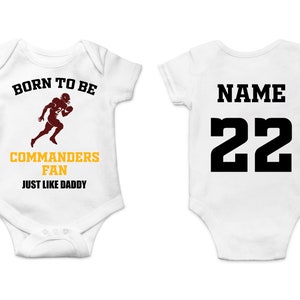 Puede incluir: Body de bebé blanco con el texto "BORN TO BE COMMANDERS FAN JUST LIKE DADDY" y un gráfico de jugador de fútbol americano granate. La parte trasera del body tiene la palabra "NAME" y el número "22" en negro.