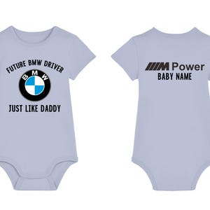 Puede incluir: Un body de bebé azul claro con un logotipo BMW negro y el texto "Future BMW Driver Just Like Daddy" en la parte delantera. La parte posterior del body tiene el texto "M Power Baby Name" en negro.