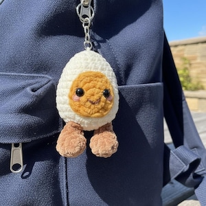 May include: A crocheted egg keychain featuring a white egg white, a golden-yellow yolk face with black safety eyes, pink embroidered cheeks, and a small brown mouth. The plush egg has two small brown feet dangling from the bottom and a silver metal chain.
