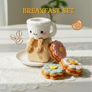May include: A crochet breakfast set featuring a white mug with a smiling face and dangling tan legs, accompanied by three flower-shaped sandwich cookies in pink, blue, and white. The text BREAKFAST SET is displayed above the items.