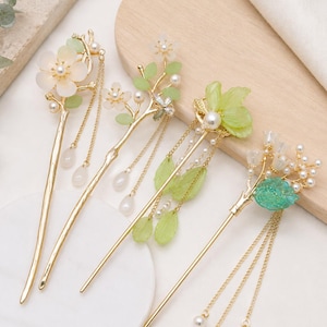 May include: Four gold-toned hair sticks featuring floral and leaf motifs in shades of green and white. Each hair accessory is adorned with small white faux pearls and delicate hanging chains with teardrop-shaped beads, creating a layered look.