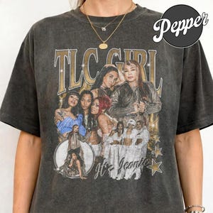 May include: A charcoal grey acid-wash t-shirt featuring a vintage-style graphic print of the music group TLC. The design includes the text "TLC GIRL" and "It's Iconic" with multiple photographic images of the group members and gold star accents.