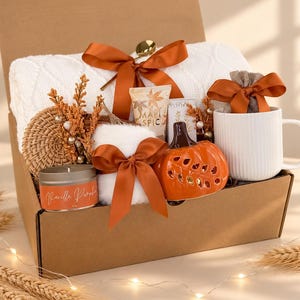 May include: A gift box containing a white cable knit blanket tied with an orange ribbon, a white ribbed mug, a 4 oz Vanilla Pumpkin candle, a ceramic orange pumpkin-shaped decor piece, a small tube of Maple Spice lotion, and a woven coaster.