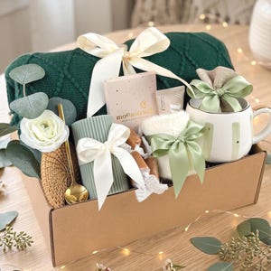Puede incluir: Una caja de regalo llena de una manta de punto verde, una taza blanca con borde dorado, una vela y otros art&iacute;culos. La caja est&aacute; atada con cintas blancas y verdes y decorada con flores y vegetaci&oacute;n. La caja est&aacute; sobre una superficie de madera.
