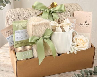 May include: A gift basket filled with a beige knit blanket, tea packets, a faux fur throw, a white mug, a candle, and a small bag tied with a bow. The basket is decorated with green ribbons and a gold spoon. The candle reads "I am Joyful".