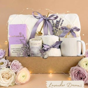 May include: A gift basket with a white blanket, a mug, a box of dark chocolate, a candle, and a small towel, all tied with lavender ribbons. The basket includes a gold spoon and lavender sprigs. The words "Lavender Dreams" are written on the bottom.
